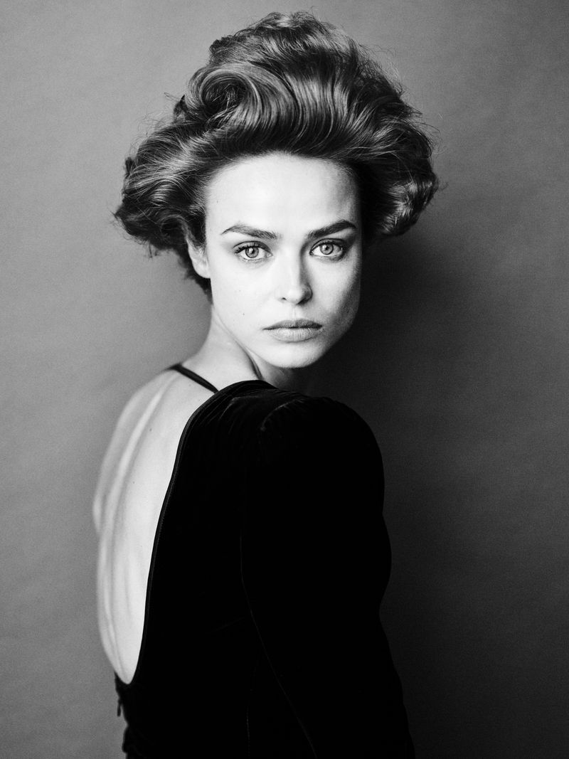 Striking black and white portrait of a woman with retro-styled hair, wearing a black velvet gown with a low-cut back, gazing directly at the camera.