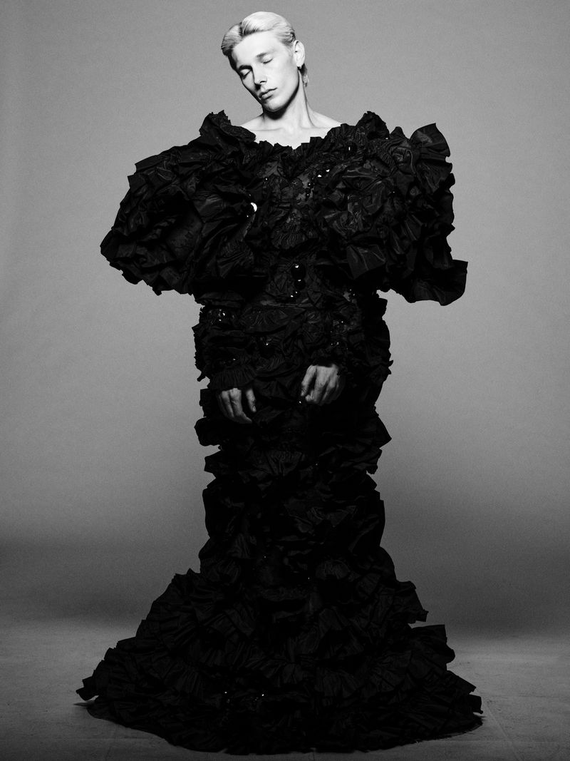 Monochrome studio shot of a slender person in a dramatic black ruffled gown with closed eyes and a serene expression on a neutral background.
