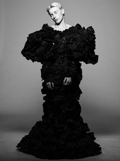 Monochrome studio shot of a slender person in a dramatic black ruffled gown with closed eyes and a serene expression on a neutral background.