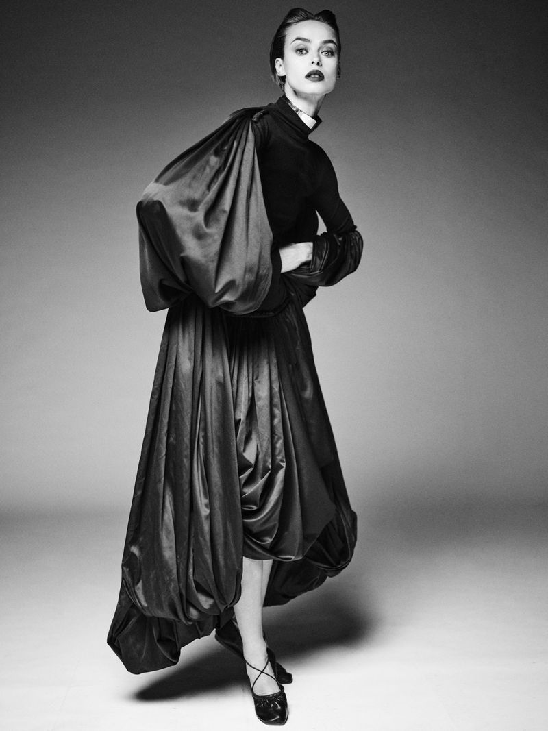 Black and white portrait of a fashion model in a voluminous skirt and top with large sleeves, posing against a neutral gradient backdrop.