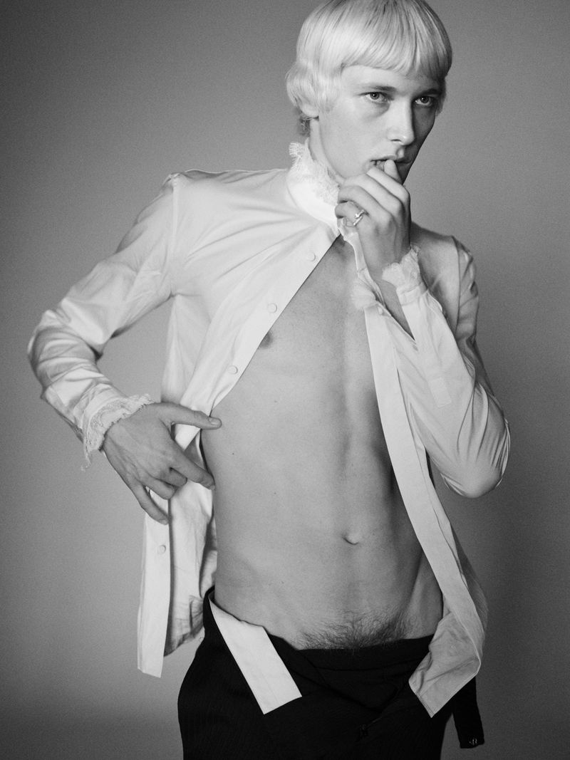 Monochrome portrait of a fair-skinned man with an unbuttoned white shirt and dark pants, posing against a gray background in the studio.