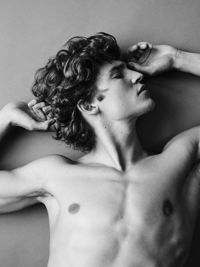 Black and white portrait of Maxime Laval shirtless with curly hair and arms raised against a neutral studio backdrop.
