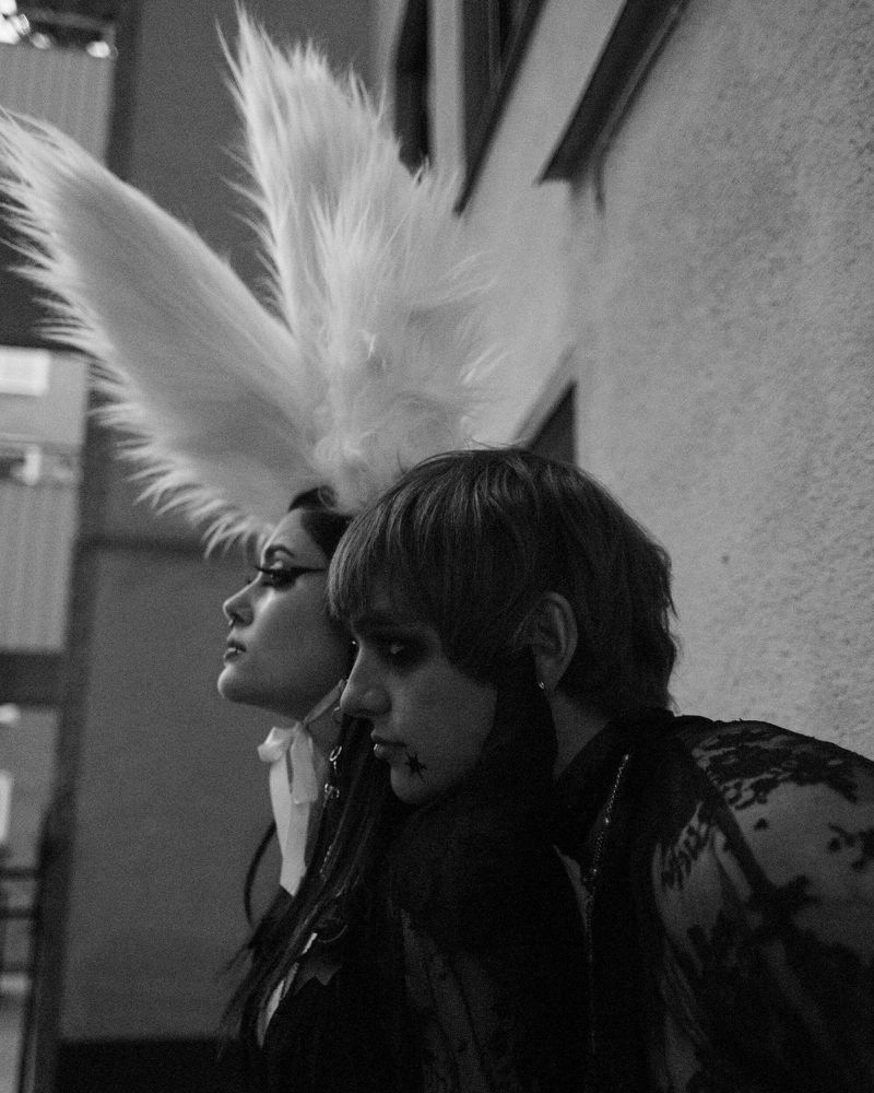 Two people pose in a black and white outdoor shot. One wears large feathered headpiece. The image has a vintage, artistic look.