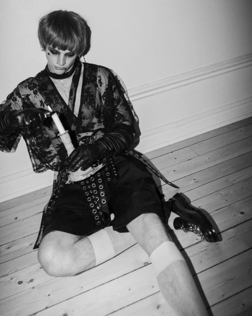 Person sits on a wood floor, lighting a candle. They wear black gloves, a lace robe, and high socks with dress shoes in a dark monochrome.