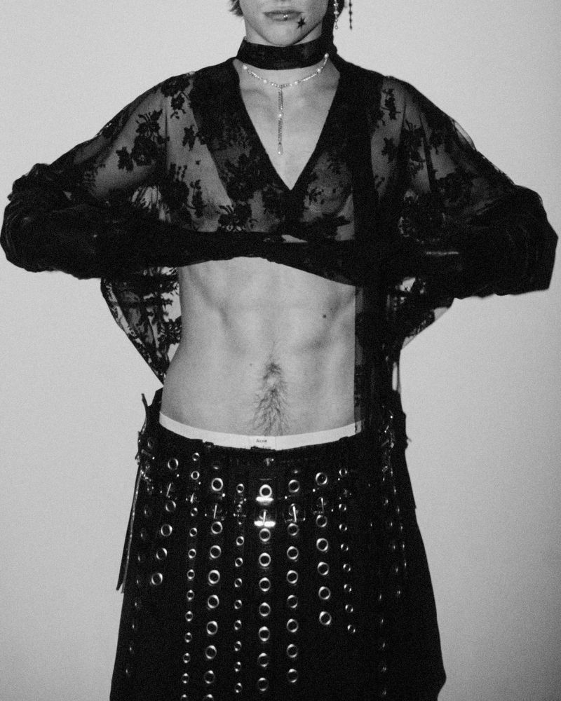 A shirtless person with visible abs wears a lace top and skirt with grommets against a solid background in a studio shot.