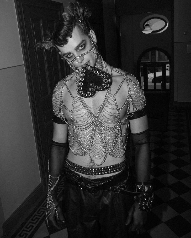 Monochrome portrait of a person with dramatic eye makeup, chains, leather clothing, and spiked accessories standing indoors on tiled floor.