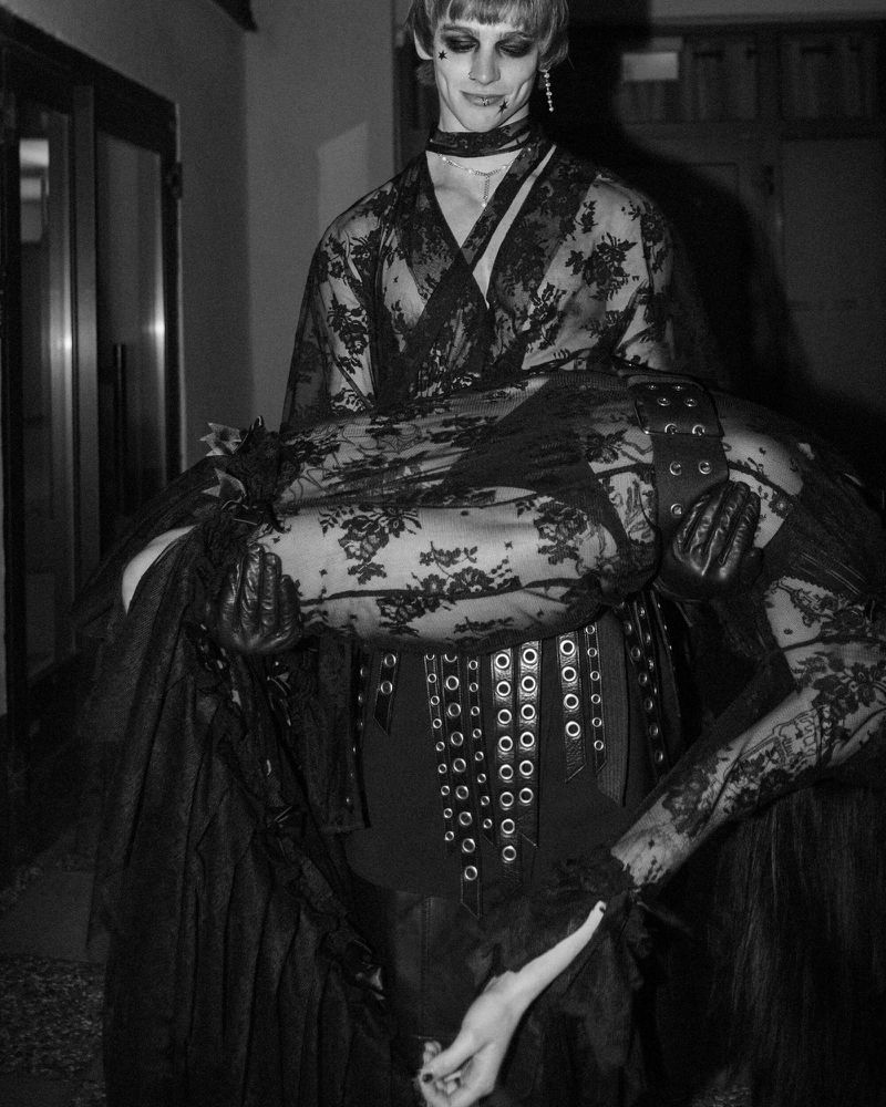 Monochrome portrait of an adult, gender-ambiguous person holding another adult in a lacey garment, dark eye makeup, and jewelry indoors.