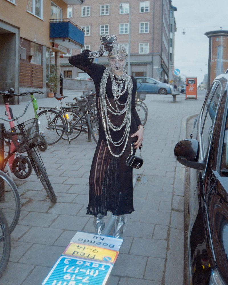 A person stands on a sidewalk next to a parked car wearing silver boots and a long black dress, posing with one arm raised near buildings.