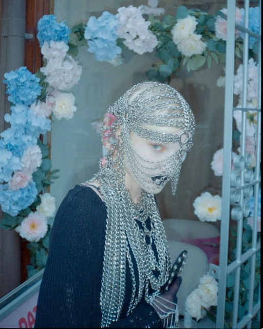 A woman with a chain headpiece is posed in front of floral decorations in an artistic fashion photograph with low-key lighting.