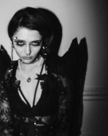Black and white portrait of a person with heavy eye makeup and pearl necklace, wearing a dark outfit with shoulder accents indoors.