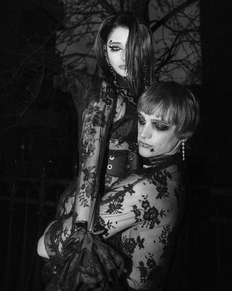 Monochrome image features two people in lace outfits against a dark background. One person carries the other. Ethereal, high-fashion.