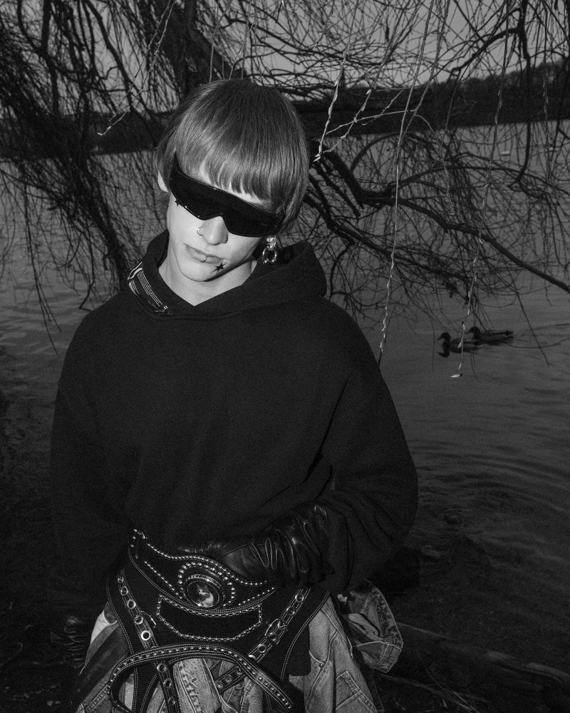 Monochrome portrait of person in sunglasses and large belt near water, with bare branches and ducks visible in the background.