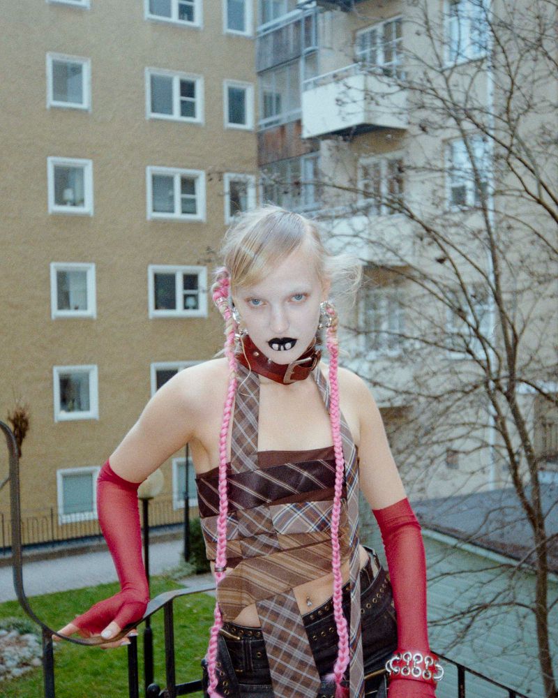 Adult woman with braided pink hair, heavy dark lipstick, and homemade top poses in front of a tall brick apartment building.