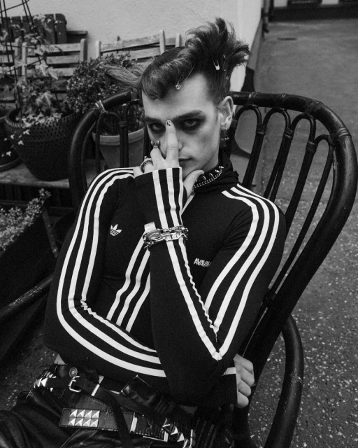 Monochrome portrait of a stylish adult male with dark eye makeup and a unique hairstyle, posing in a black and white setting with greenery.