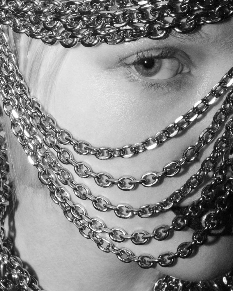Black and white close-up of a person's eye and face partially covered by draped metal chains creating a veiled effect with artistic feel.