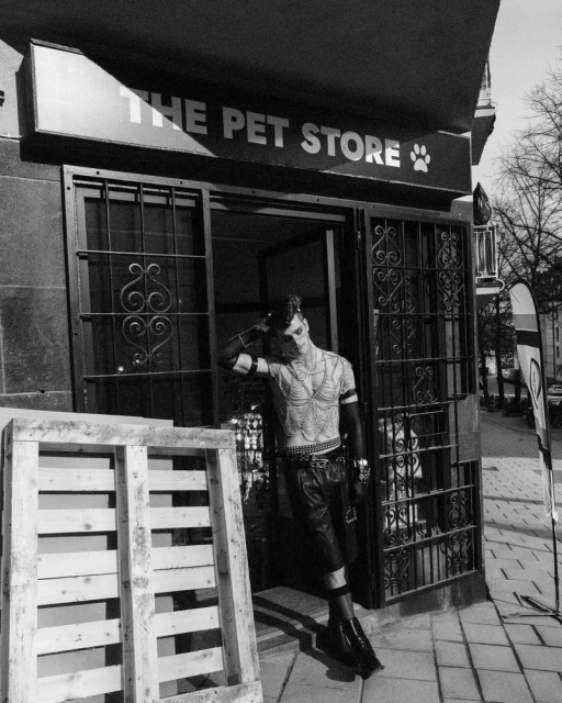 Monochrome portrait of tattooed man in leather shorts leaning against a pet store entrance with a wooden pallet in the foreground.