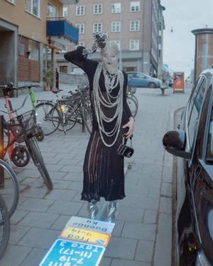 Fashion model in a chainmail mask, black knit dress, draped silver chains, and metallic boots on a city street.