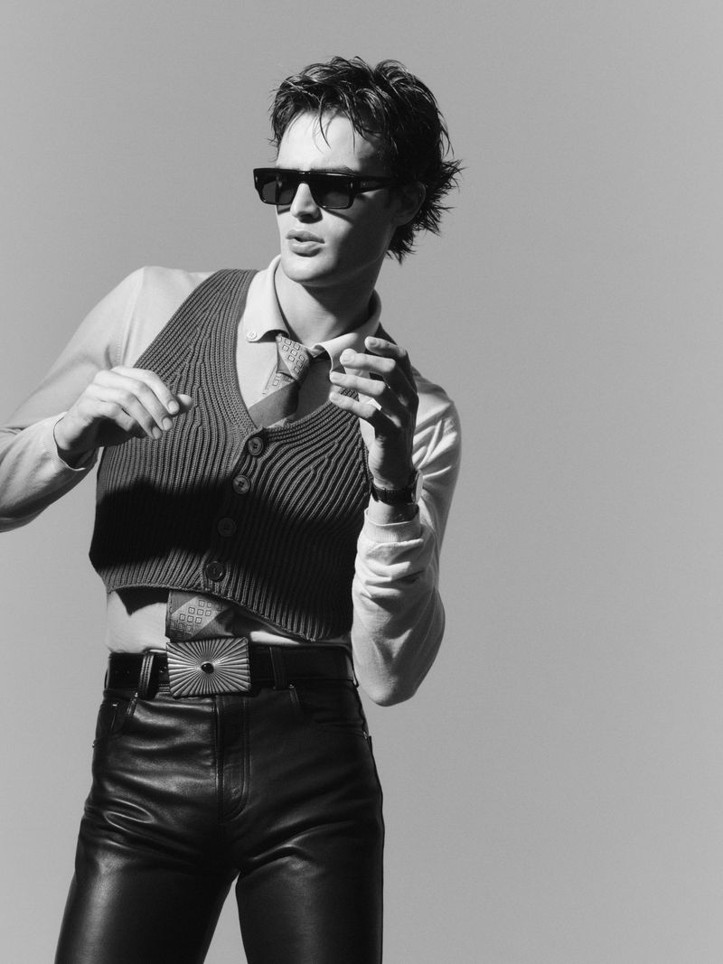 A stylish adult male with dark hair, wearing sunglasses, a vest, and leather pants poses against a neutral background in black and white.