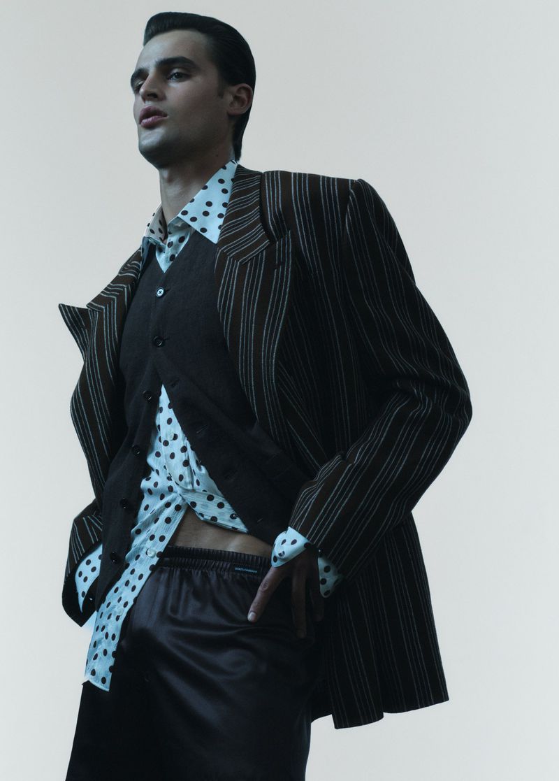 A male fashion model poses in a pinstripe suit jacket, button-down vest, spotted shirt, and dark shorts against an off-white background.