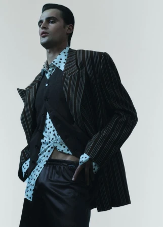 A male fashion model poses in a pinstripe suit jacket, button-down vest, spotted shirt, and dark shorts against an off-white background.