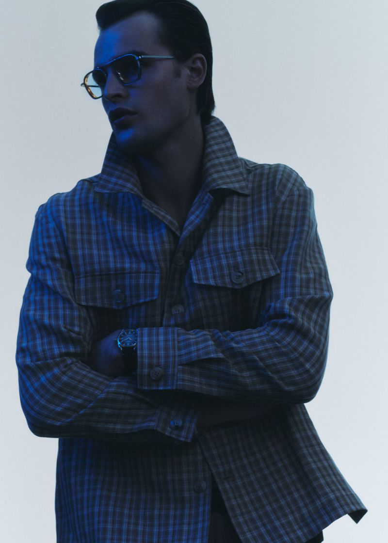 Stylish man in eyewear and a patterned shirt with folded arms, posing in a studio with soft lighting for a fashion editorial.