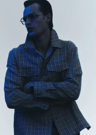 Stylish man in eyewear and a patterned shirt with folded arms, posing in a studio with soft lighting for a fashion editorial.