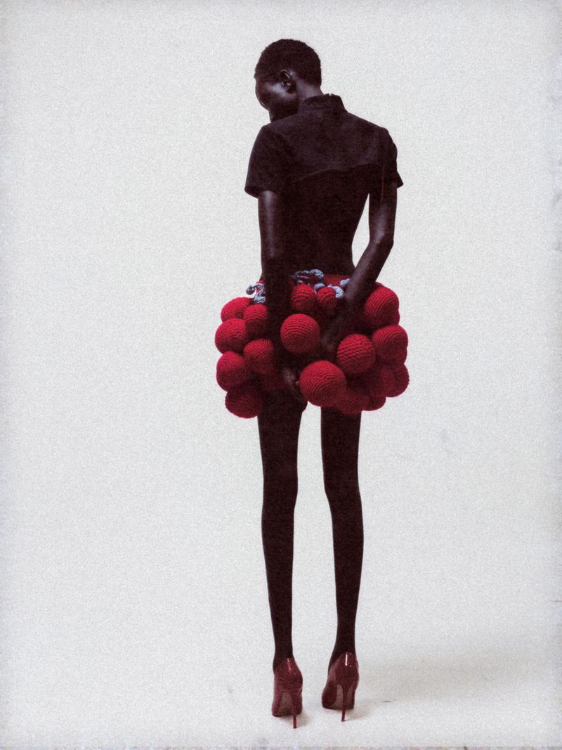 Fashion shot of a slender dark-skinned model wearing a unique ball-shaped skirt and heels, posing in a bright white studio setting.