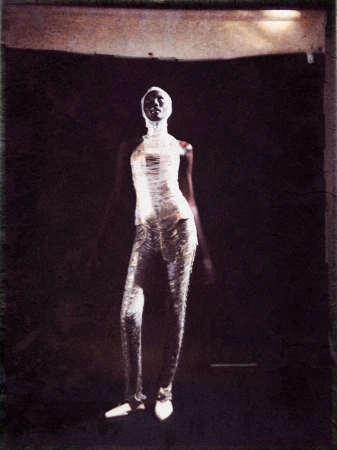 Fashion shot of an African American woman in a light-colored, form-fitting outfit, head wrap, and low heels against a dark background.