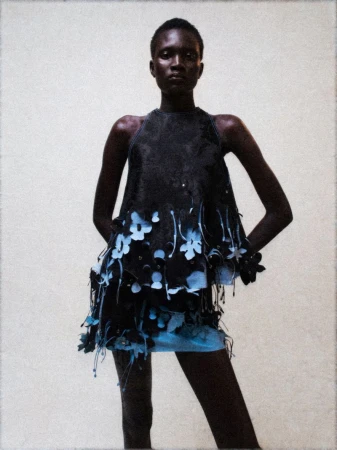 Black female model poses in dark top and light blue skirt with flower detail against a neutral backdrop in a studio setting.