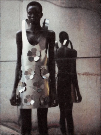 Editorial shot of a young Black woman in a metallic dress with flower accents, mirrored by a figure in the background against a wall.