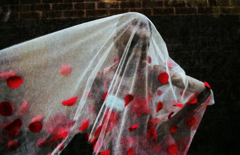 Person shrouded in a sheer white fabric, adorned with scattered red flower petals, standing against a dark brick wall background.