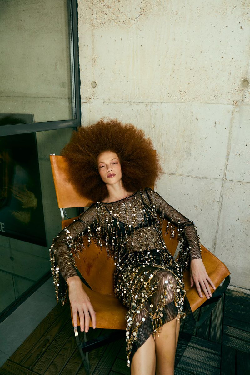 Woman with large afro sits in chair wearing a see-through black dress with gold sequins against concrete and a window backdrop.