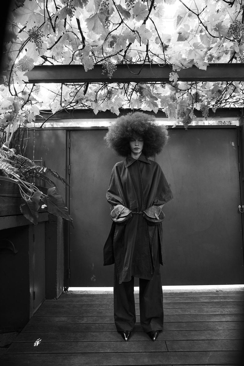 A woman with an afro stands in front of a gray door surrounded by vegetation in this black and white fashion photograph.