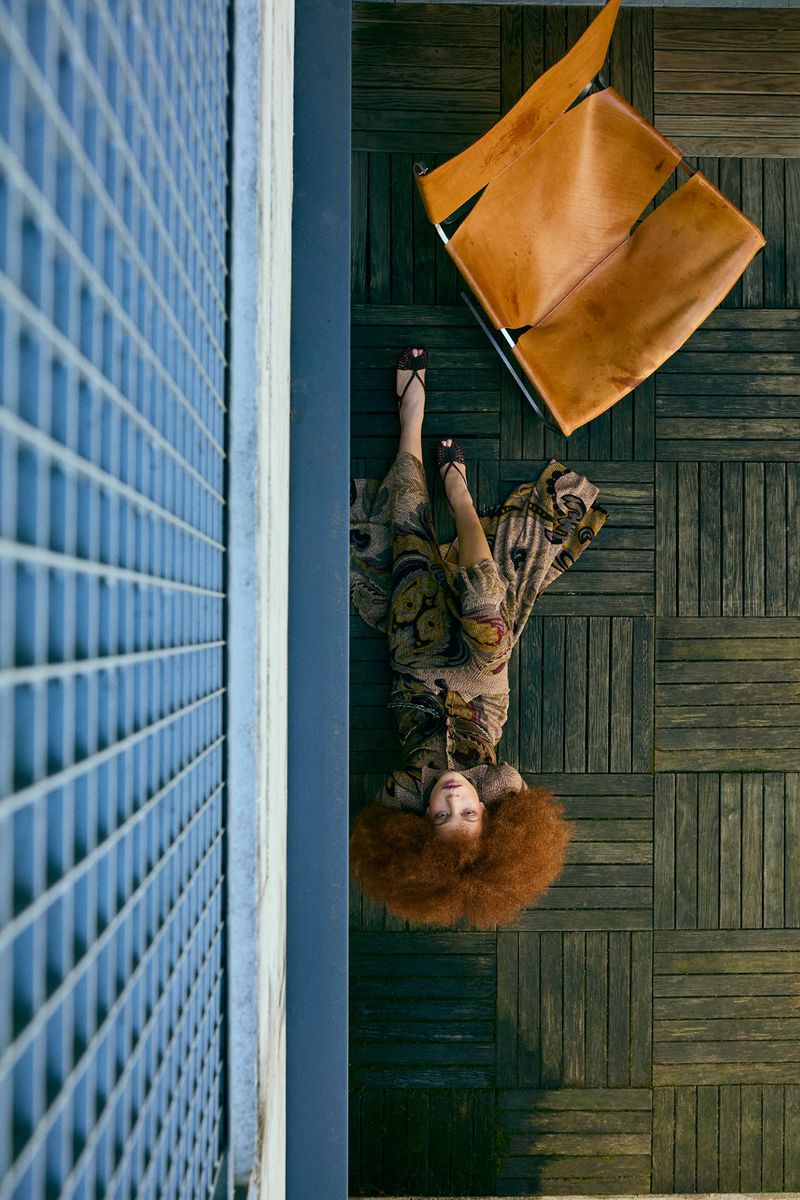 Overhead shot of an adult woman with auburn hair lying on a wooden floor near a mid-century modern leather chair and a blue wall with a grid.