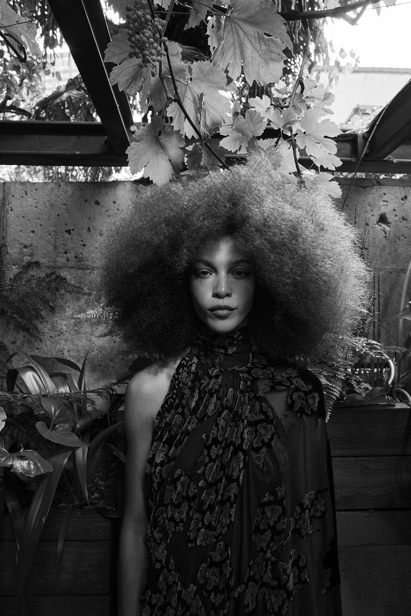 Model with voluminous afro hairstyle wearing a patterned high-neck top outdoors surrounded by leafy vines and foliage.