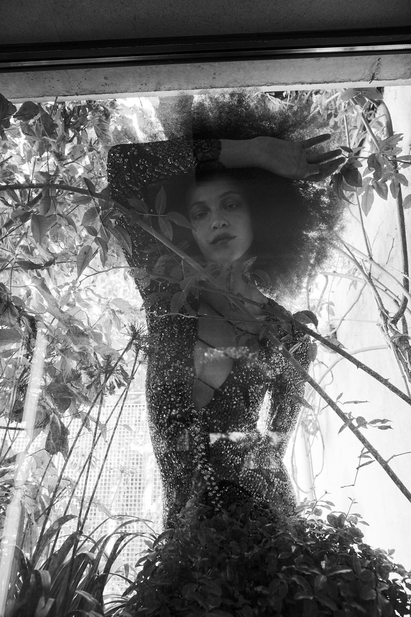 Black and white portrait of a woman with an afro wearing a sparkly dress, standing indoors amid lush green plants.