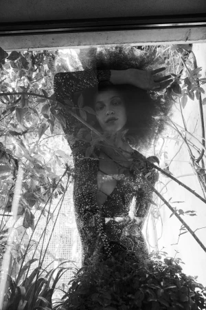 Black and white portrait of a woman with an afro wearing a sparkly dress, standing indoors amid lush green plants.