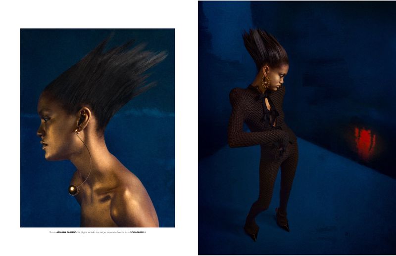 Fashion editorial portrait of a Black woman with a gravity-defying hairstyle, elaborate gold jewelry, and shimmering skin tone on a blue backdrop.