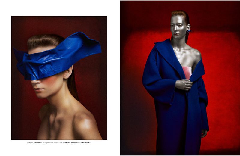 Conceptual fashion shot featuring a woman with a large blue fabric mask and a woman with metallic body paint in a blue coat.