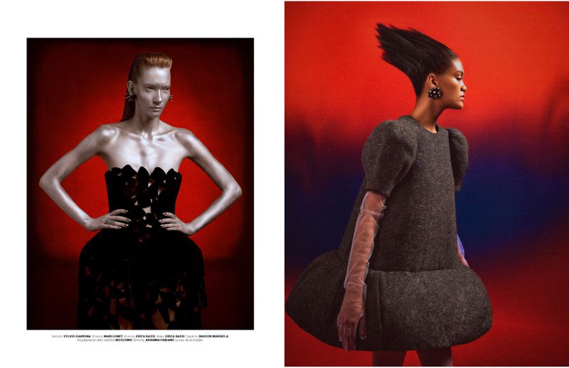 Two women models wearing dark haute-couture gowns, posing indoors against a vibrant red gradient backdrop in a fashion editorial shot.