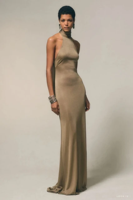 Elegant woman in a floor-length halter dress posing against a plain studio backdrop, showcasing the garment's shimmering fabric and cut.