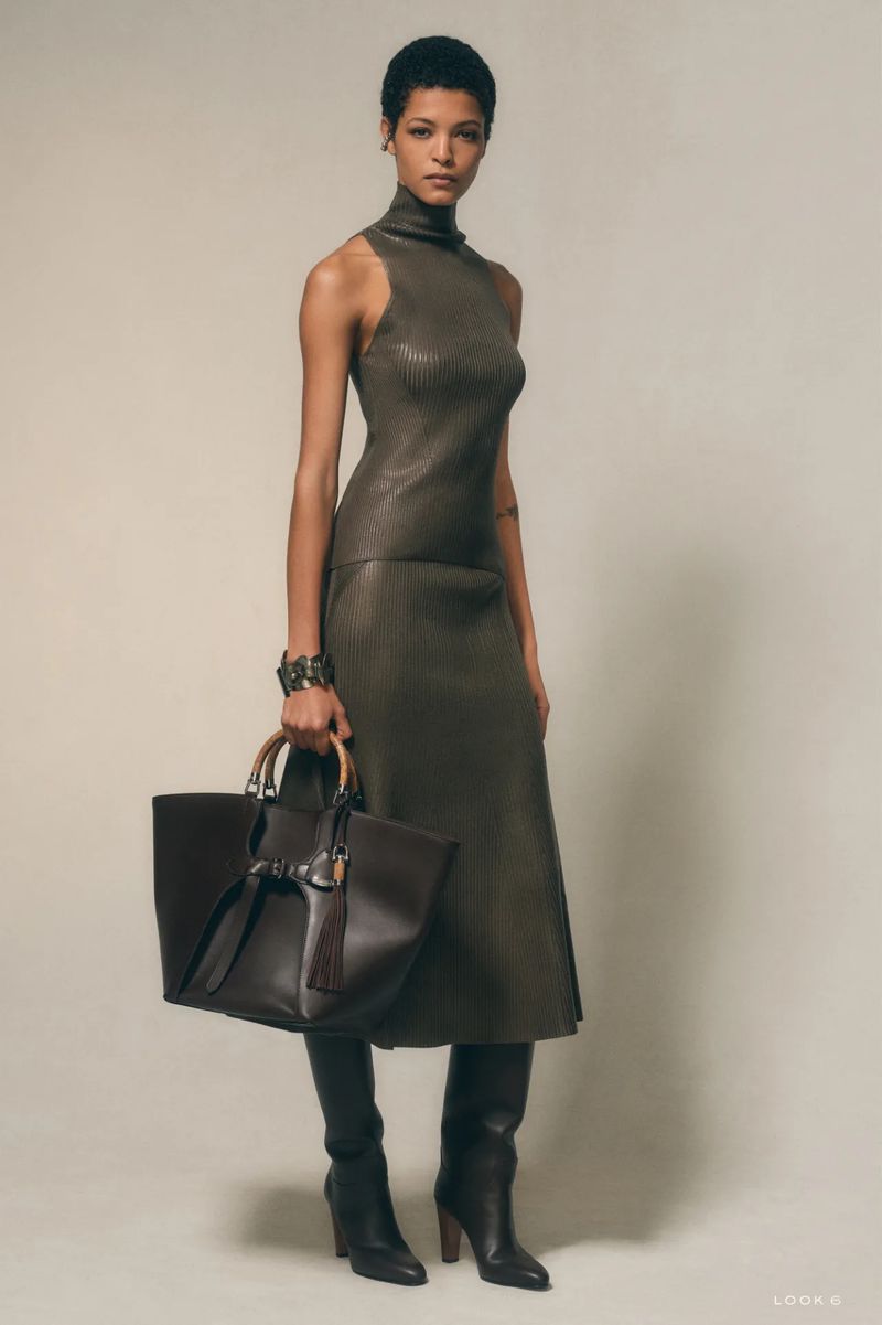 Fashion editorial shot of a woman posing in an olive-green dress, tall boots, and holding a dark brown leather tote bag.