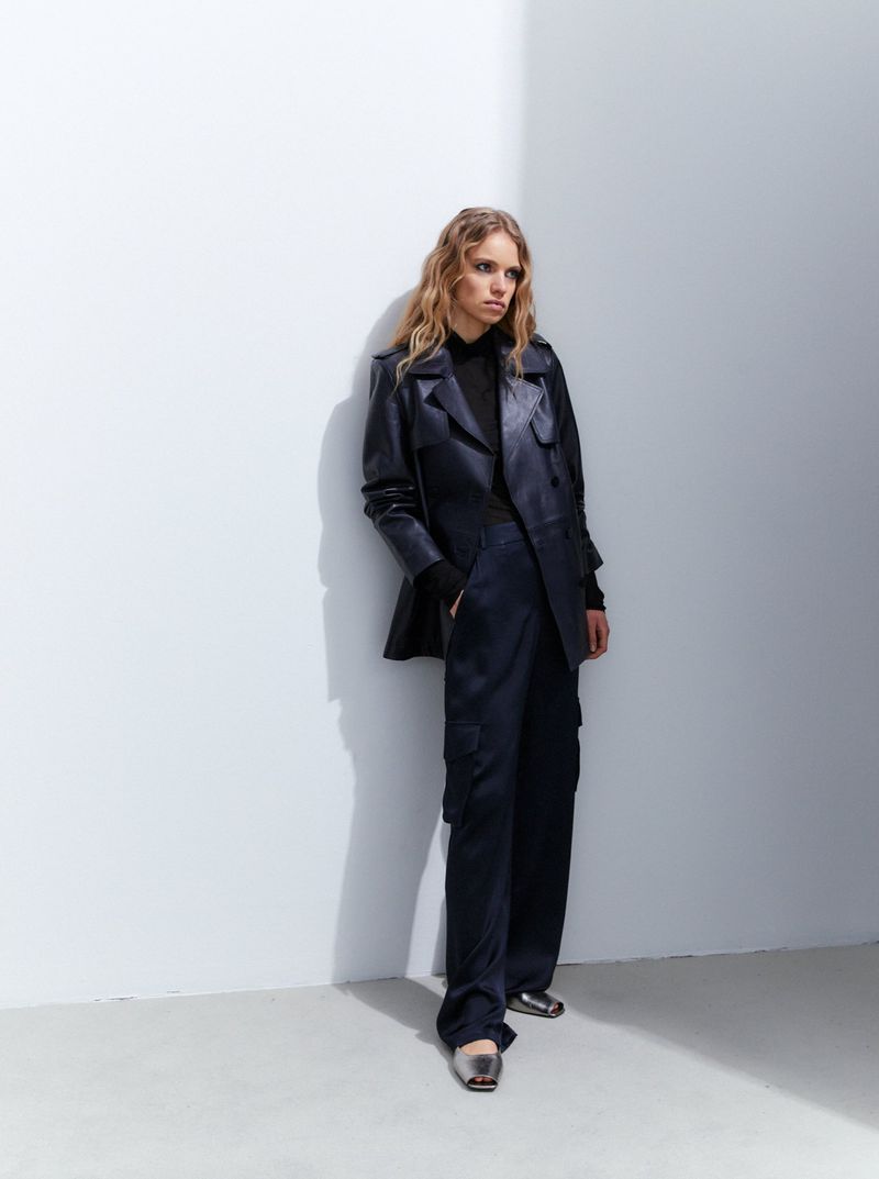 A fashionable adult woman poses in a navy blue suit against a white wall, showcasing modern sophistication in a studio setting.