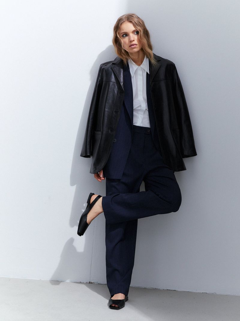 A woman in a black blazer, white shirt, and dark pants poses against a white wall, looking off to the side in a fashion editorial style.