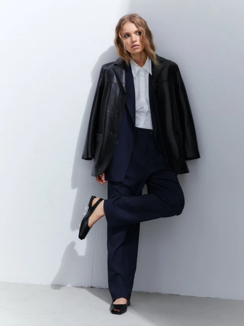 A woman in a black blazer, white shirt, and dark pants poses against a white wall, looking off to the side in a fashion editorial style.