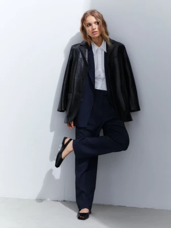 A woman in a black blazer, white shirt, and dark pants poses against a white wall, looking off to the side in a fashion editorial style.