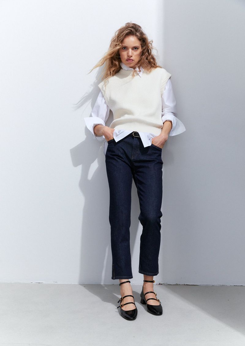 A woman stands in front of a white wall wearing a white vest, button-down shirt, dark jeans, and black flats with ankle straps.