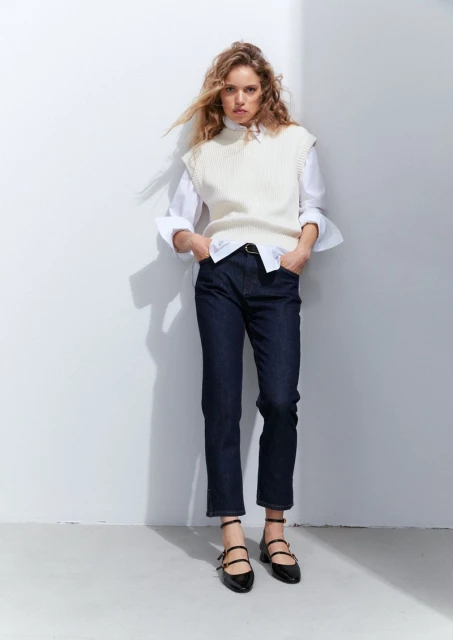 A woman stands in front of a white wall wearing a white vest, button-down shirt, dark jeans, and black flats with ankle straps.