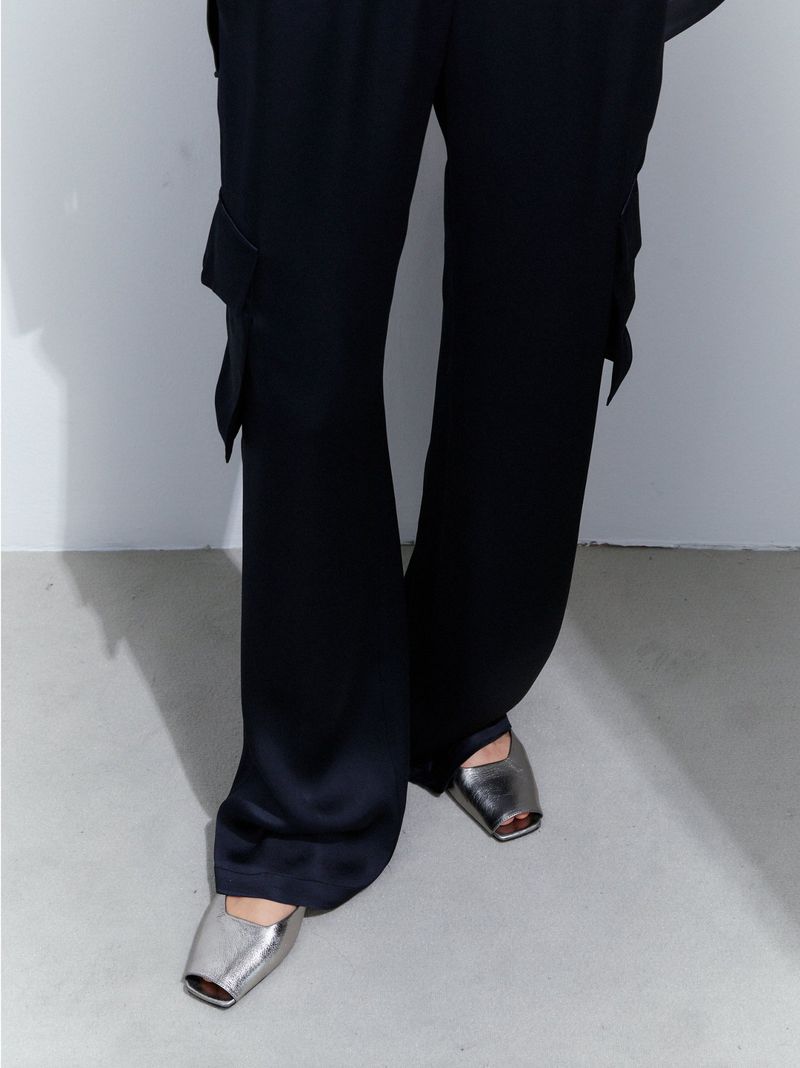 Close-up of a person wearing black satin pants with cargo pockets and silver metallic square-toe shoes, captured in a studio.