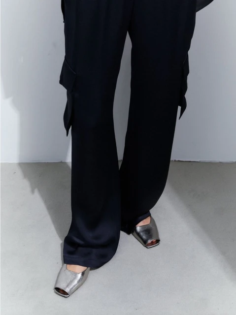 Close-up of a person wearing black satin pants with cargo pockets and silver metallic square-toe shoes, captured in a studio.